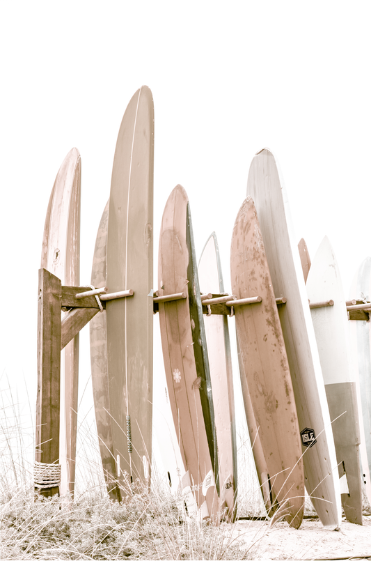Best-selling soft neutral surfboard fine art print, perfect for coastal home decor and beach house interiors. Minimalist vertical surf art print showcasing a line-up of surfboards in various lengths and soft, neutral pastel hues, resting on a simple wooden rack with some dry grass in the foreground. This piece of surf wall decor features a bright, hazy white backdrop, enhancing its clean aesthetic. Perfect as pastel surf decor or for adding surf vibes to kids surf room decor or a beach house.