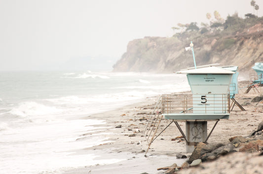 Photography by Kim Dybczak of: A light blue lifeguard tower stands on wet sand with gentle waves nearby.