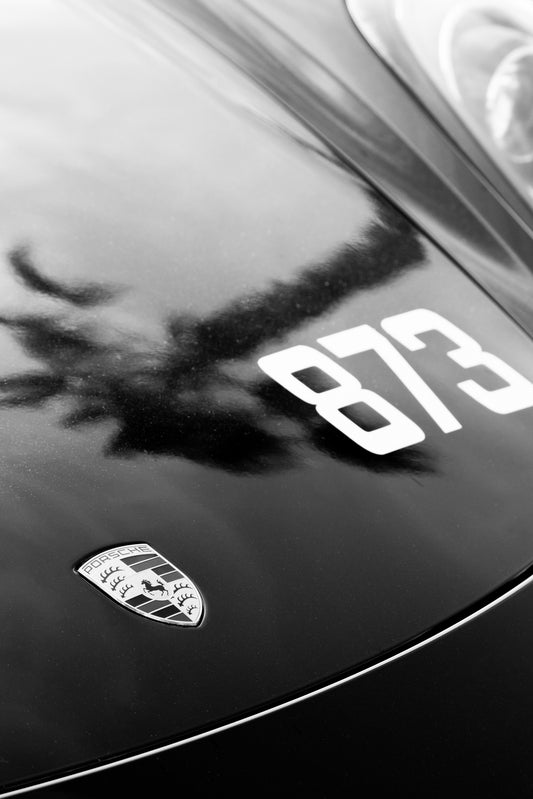Photography by Salt Creek Prints of: Porsche 8312, Close up black and white image of the hood of a modern Porsche with a Palm Tree reflected on the hood.