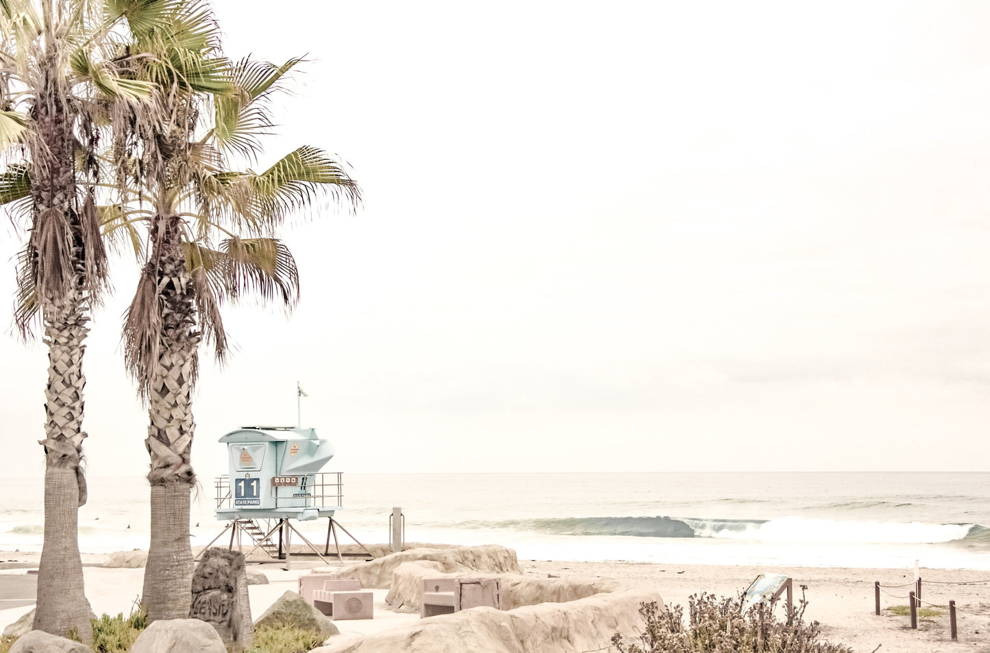 Photography by Kim Dybczak of: Two tall palms and a pastel blue lifeguard tower set a calm, dreamy beach scene.