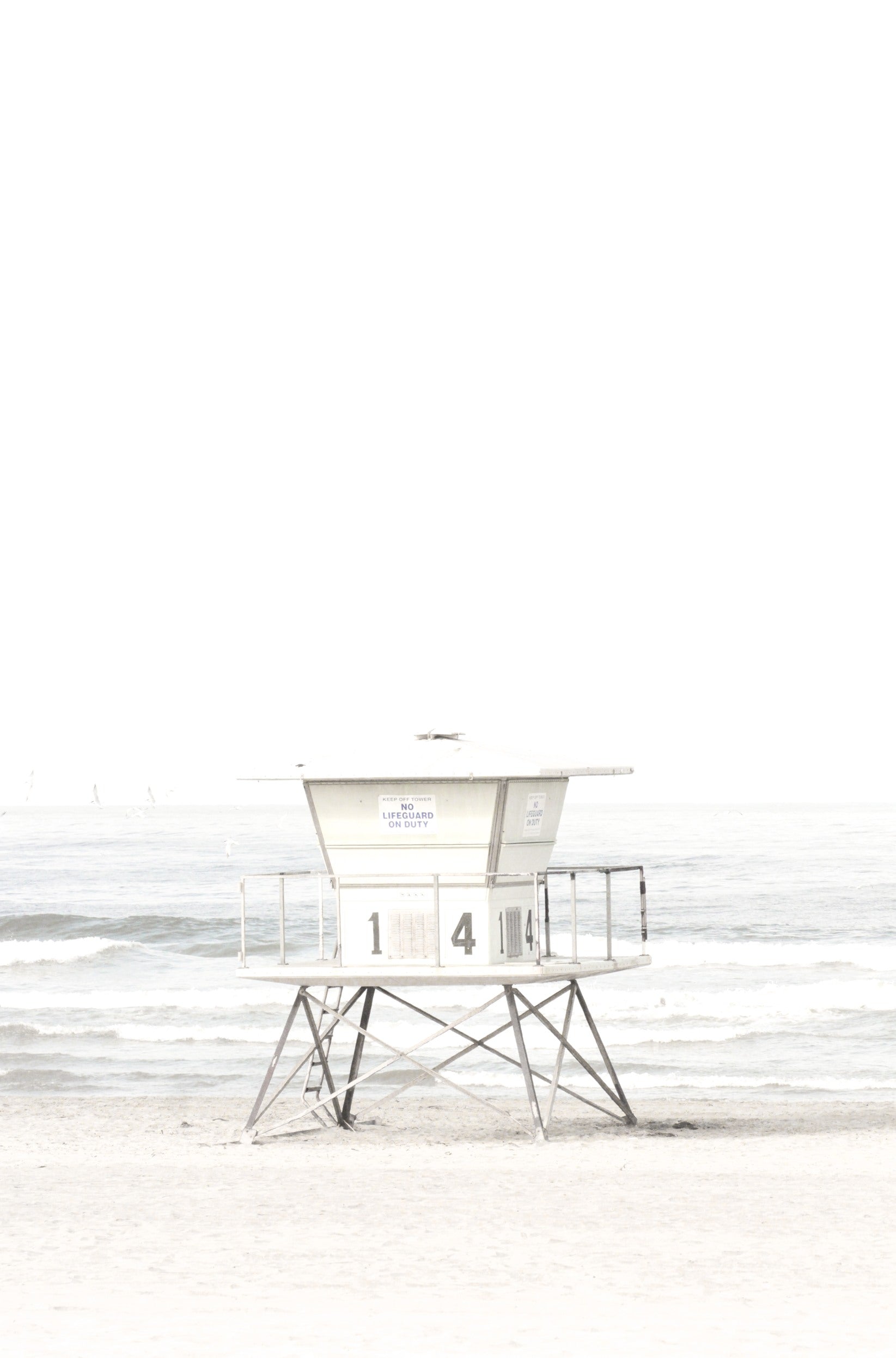 Photography by Kim Dybczak of: A pale lifeguard tower stands near the shore with ocean waves in the distance.