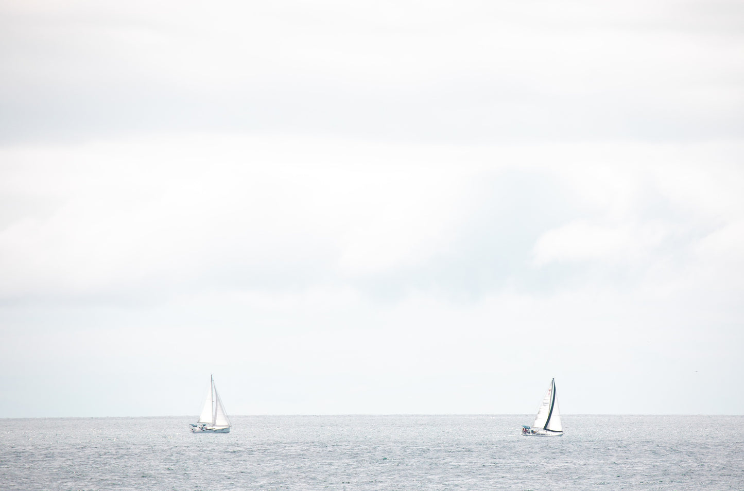 Photography by Kim Dybczak of: A single sailboat moves across the water off the coast, with a soft horizon beyond.