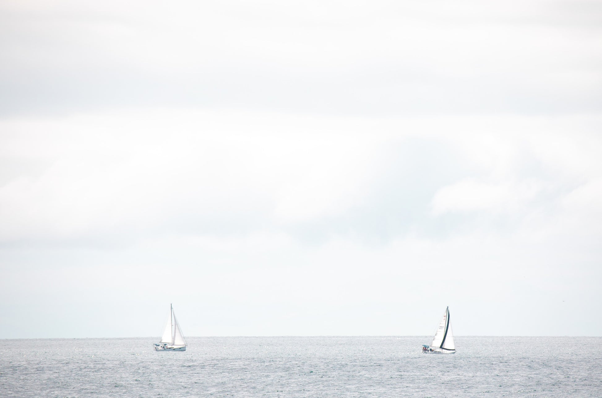 Photography by Kim Dybczak of: A single sailboat moves across the water off the coast, with a soft horizon beyond.