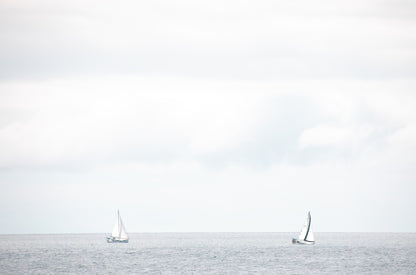 Photography by Kim Dybczak of: A single sailboat moves across the water off the coast, with a soft horizon beyond.