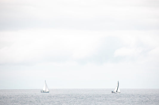 Photography by Kim Dybczak of: A single sailboat moves across the water off the coast, with a soft horizon beyond.
