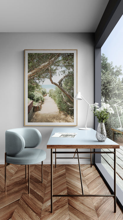 A serene landscape coastal photography print showing a path leading down to the ocean, framed by lush coastal foliage and winding branches. This vertical coastal photography piece captures the beauty of the Seaside Beach, on the California coast, perfect for a beach house decor. Photograph in minimalist mock up.