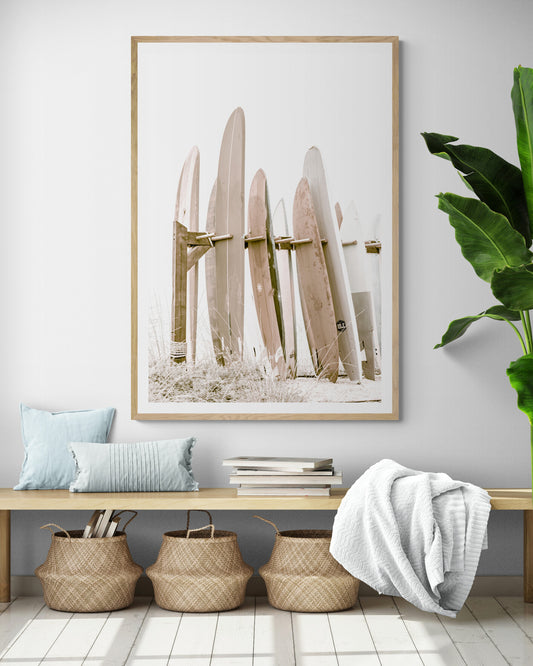 Framed neutral-toned surfboard wall art, ideal for modern, minimalist, and beach-style homes. Minimalist vertical surf art print showcasing a line-up of surfboards in various lengths and soft, neutral pastel hues, resting on a simple wooden rack with some dry grass in the foreground. This piece of surf wall decor features a bright, hazy white backdrop, enhancing its clean aesthetic. Perfect as pastel surf decor or for adding surf vibes to a beach house. Photograph in entry way mock up