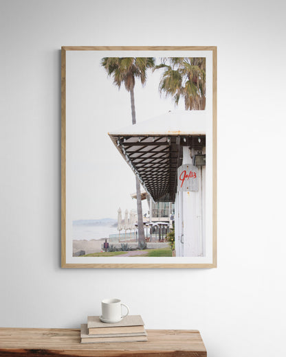 A framed beach scene art print with a wooden structure and palm trees, titled 'Jake's' on the sign, set against a background of a beach and ocean. Photograph in minimalist mock up.