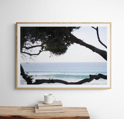 An ocean view reveals surfers on gentle waves, with a white bird soaring. Tree branches provide a natural, rustic foreground to the serene scene, taken at a beach in Del Mar, San Diego. Photograph in minimalist mock up.