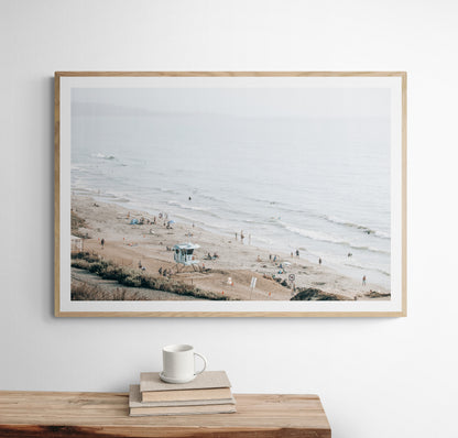A slightly hazy, high-angle Landscape Beach Print captures a wide beachscape print where a light blue ocean waves meet a sandy shore dotted with numerous people enjoying the Summer Vibes. Gentle white waves roll towards the beach. A Blue Lifeguard Tower stands prominently in the lower center of the framed landscape beach print. The overall lighting suggests a bright, overcast travel photography day on the San Diego coast. Photograph in minimalist mock up
