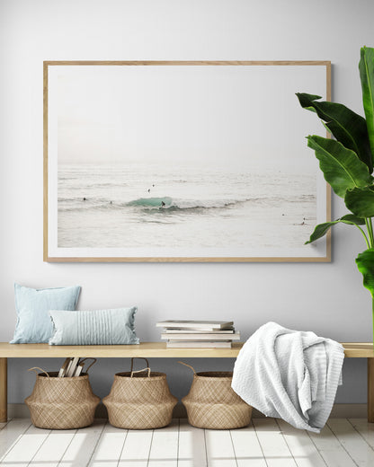 Fine art of horizontal landscape print by Southern California photographer Kim Dybczak of Salt Creek Prints, featuring a bright image captures a pale sandy beach giving way to a vast, light gray ocean with gentle waves. Several surfers are scattered across the water, some sitting on their boards and others riding small waves. The sky is a uniform, pale white, creating a soft, overcast atmosphere, located in Windansea Beach in La Jolla, California. Photograph in framed minimalist entry way mock up