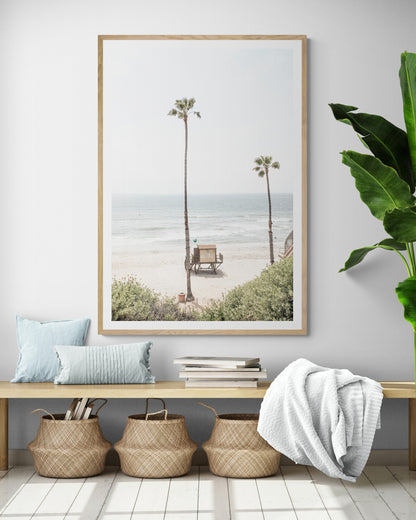 Vertical beach wall art featuring a lifeguard tower nestled between two tall palm trees on a sunny California beach, capturing a serene beach vibe. Photograph in minimalist mock up.