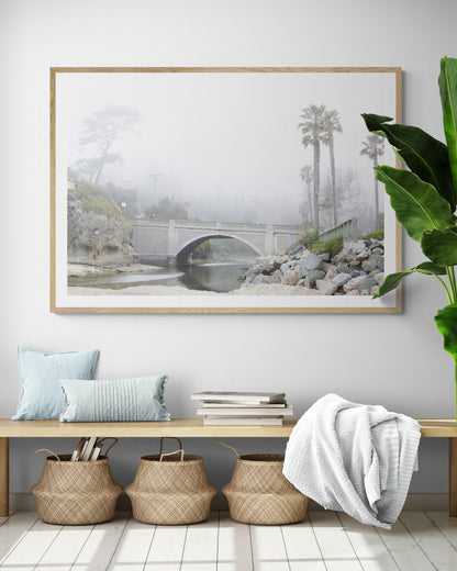 Landscape coastal photography of a concrete arch bridge disappearing into a dense fog, flanked by palm trees and rocky shores. This calm beach print evokes a mysterious seascape. Photograph in minimalist mock up.