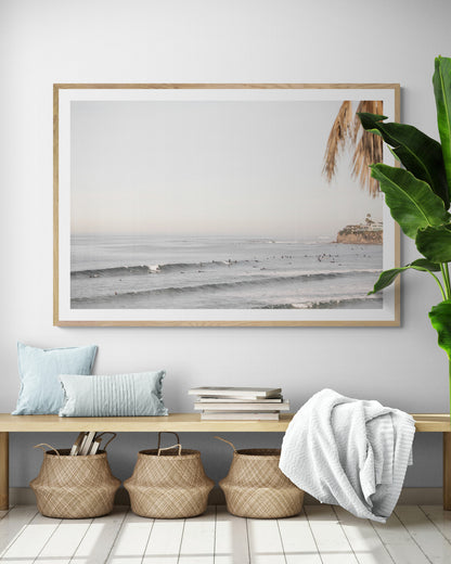 Landscape beach photography of Tourmaline Beach, Pacific Beach, California, with waves and many surfers, framed by a palm tree in the corner. This ocean print captures casual surf vibes. Photograph in minimalist mock up.