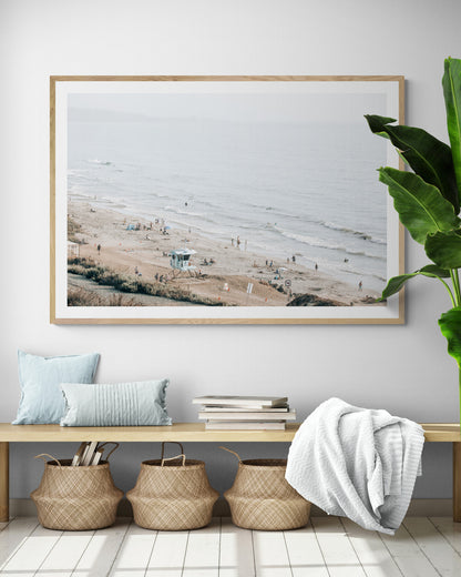 A slightly hazy, high-angle Landscape Beach Print captures a wide beachscape print where a light blue ocean waves meet a sandy shore dotted with numerous people enjoying the Summer Vibes. Gentle white waves roll towards the beach. A Blue Lifeguard Tower stands prominently in the lower center of the framed landscape beach print. The overall lighting suggests a bright, overcast travel photography day on the San Diego coast. Photograph in minimalist mock up