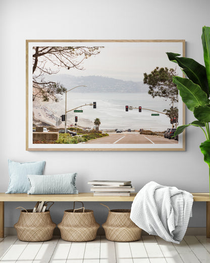 A hazy eye-level shot of San Diego Streets looks down a sloping road featuring red traffic lights and "Carmel Valley" signs, capturing a Local Surf Print vibe. The foreground of this California Wall Art showcases landscape and cars, framed by tree branches at the top, adding to the San Diego Photo aesthetic. The lighting suggests a daytime, perfect for Minimalist Beach Wall Art. This horizontal San Diego print makes a great piece of horizontal beach wall decor. Photograph in minimalist entry way mock up