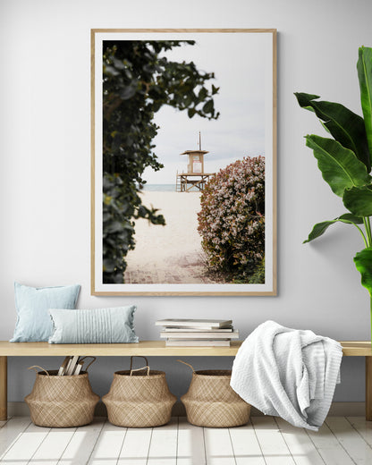 Vertical coastal photography of a Newport Beach lifeguard tower on the sand, framed by lush green and flowering foliage. Photograph in minimalist mock up.