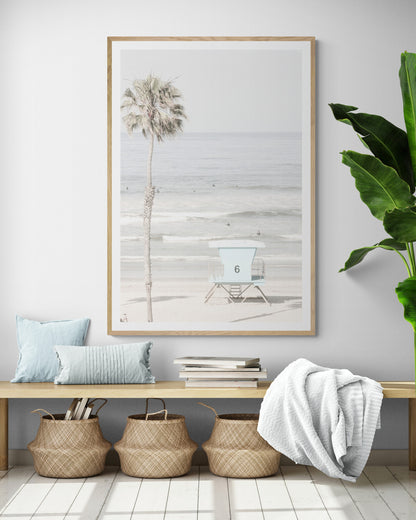 Dreamy coastal palm tree print featuring a tall palm tree with shaggy trunks in the foreground and a pastel blue lifeguard tower centered in the distance. This vertical tropical print is captured at Oceanside Beach on the Southern California coast, is bathed in bright, hazy sunlight, creating a pastel beach art effect. Photograph in minimalist mock up.