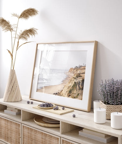 Fine art of Torrey Pines, San Diego California horizontal print of hazy high-angle landscape beach photography captures a light blue ocean meeting a sandy beach with many beach goers. To the right, tall tan and brown cliffs rise as iconic coastal scenery. Photograph in minimalist framed mock up.