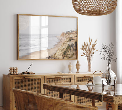 Fine art of Torrey Pines, San Diego California horizontal print of hazy high-angle landscape beach photography captures a light blue ocean meeting a sandy beach with many beach goers. To the right, tall tan and brown cliffs rise as iconic coastal scenery. Photograph in minimalist framed dining room mock up.