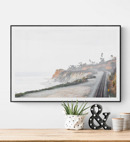 A slightly hazy, wide Landscape Coastal Photography shot shows a set of train tracks curving along a coastal cliffside, San Diego Coast. To the left, a sandy beach meets a light blue ocean waves with gentle waves, where a few small figures of people are visible. The cliffs are varied in color, with sections of tan, white, and hints of orange, topped with greenery and a few Palm Trees. The sky is a muted white, perfect for Minimalist Surf Wall Art. Photograph in minimalist mock up