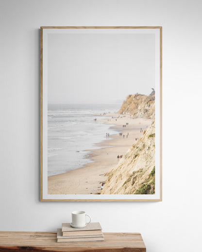 Fine art of Torrey Pines, San Diego California vertical print of hazy high-angle landscape beach photography captures a light blue ocean meeting a sandy beach with many beach goers. To the right, tall tan and brown cliffs rise as iconic coastal scenery. Photograph in minimalist framed mock up.