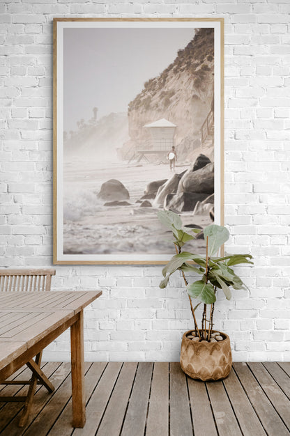 Vertical surf art print of a lone surfer carrying a surfboard, walking on a rocky, foggy shoreline at Moonlight State Beach with a lifeguard tower and cliff in the background. Photograph in minimalist mock up.