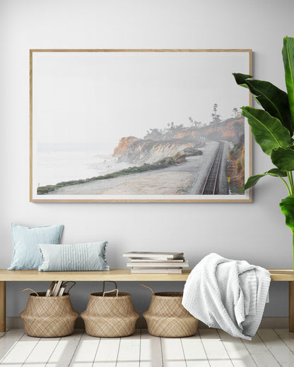 A slightly hazy, wide Landscape Coastal Photography shot shows a set of train tracks curving along a coastal cliffside, San Diego Coast. To the left, a sandy beach meets a light blue ocean waves with gentle waves, where a few small figures of people are visible. The cliffs are varied in color, with sections of tan, white, and hints of orange, topped with greenery and a few Palm Trees. The sky is a muted white, perfect for Minimalist Surf Wall Art. Photograph in minimalist entry way mock up