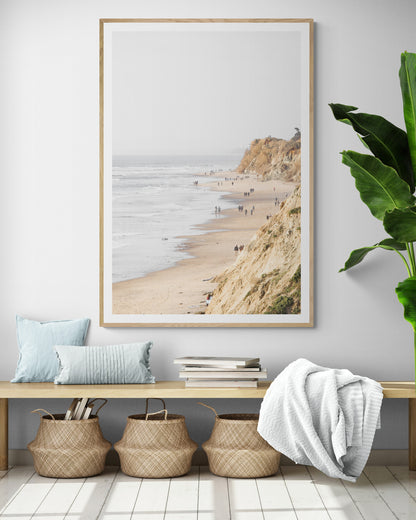 Fine art of Torrey Pines, San Diego California vertical print of hazy high-angle landscape beach photography captures a light blue ocean meeting a sandy beach with many beach goers. To the right, tall tan and brown cliffs rise as iconic coastal scenery. Photograph in minimalist framed entry way mock up.