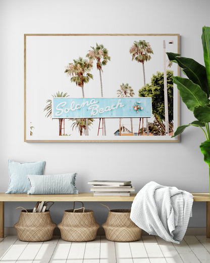 Colorful travel photography capturing the iconic 'Solana Beach' sign, a quintessential piece of Surf Signs San Diego art. The retro light blue sign, adorned with white cursive script and a unique steer head logo, is set against a backdrop of towering palm trees and a bright, hazy California sky. This Southern California surf beach sign evokes strong summer vibes and is perfect for coastal decor. Photograph in minimalist framed entry way mock up.