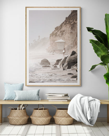 Vertical surf art print of a lone surfer carrying a surfboard, walking on a rocky, foggy shoreline at Moonlight State Beach with a lifeguard tower and cliff in the background. Photograph in minimalist mock up.